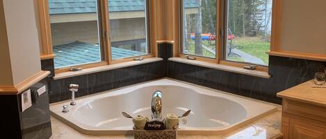 A bathroom with a large freestanding bathtub, a vanity with a mirror, and a window with a view of trees and a lake.