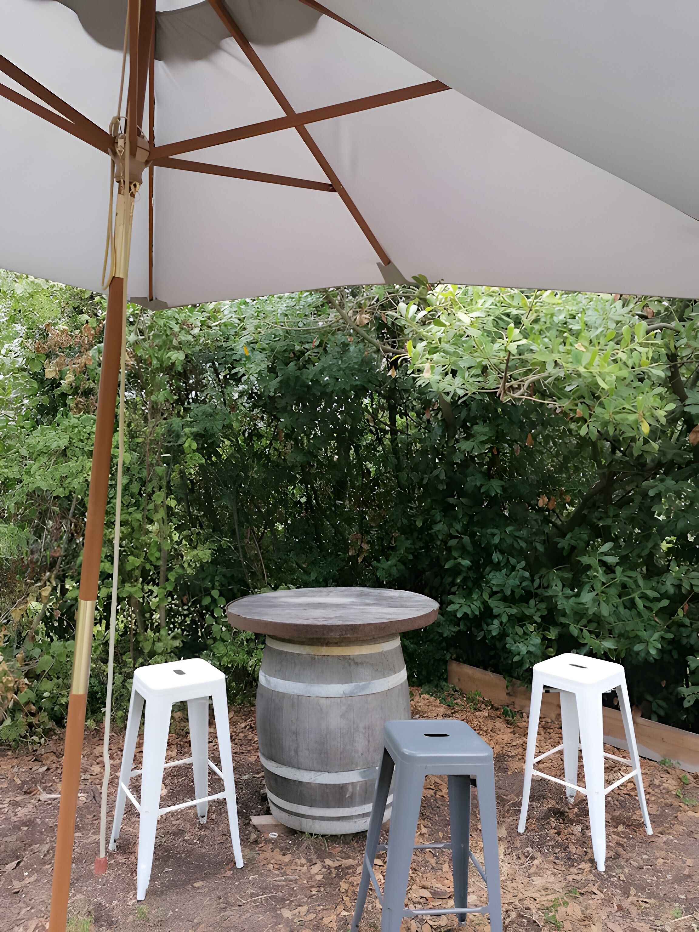 A round table with two white stools, a large barrel, and a white umbrella in an outdoor setting with dense foliage.