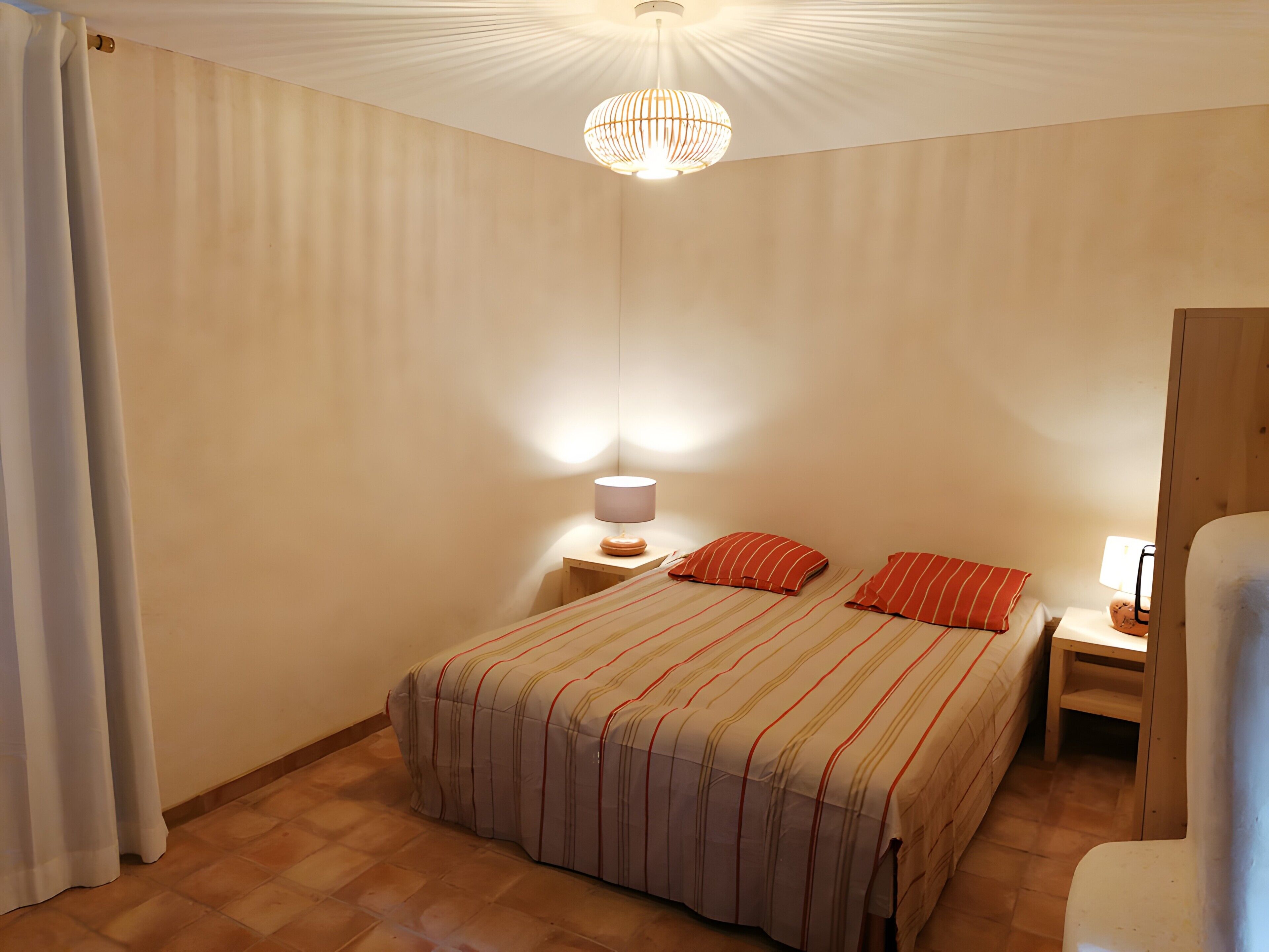 A bedroom with a bed, two nightstands, a lamp, a hanging light fixture, and a wooden door.