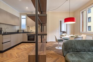 A modern kitchen with wooden flooring, a dining area with a red pendant light, and a view of the outside through a window.