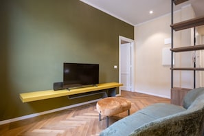 A modern living room with a flat-screen TV on a yellow stand, a herringbone-patterned wooden floor, a grey sofa, and a small round ottoman. - Luxury Bridge Apartment (Florence)