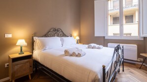 Uma cama bem arrumada com lençóis brancos e toalhas enroladas, uma mesa de cabeceira com abajur e uma janela com vista para uma varanda.