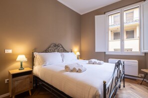 A neatly made bed with white linens and rolled towels, a bedside table with a lamp, and a window with a view of a balcony.