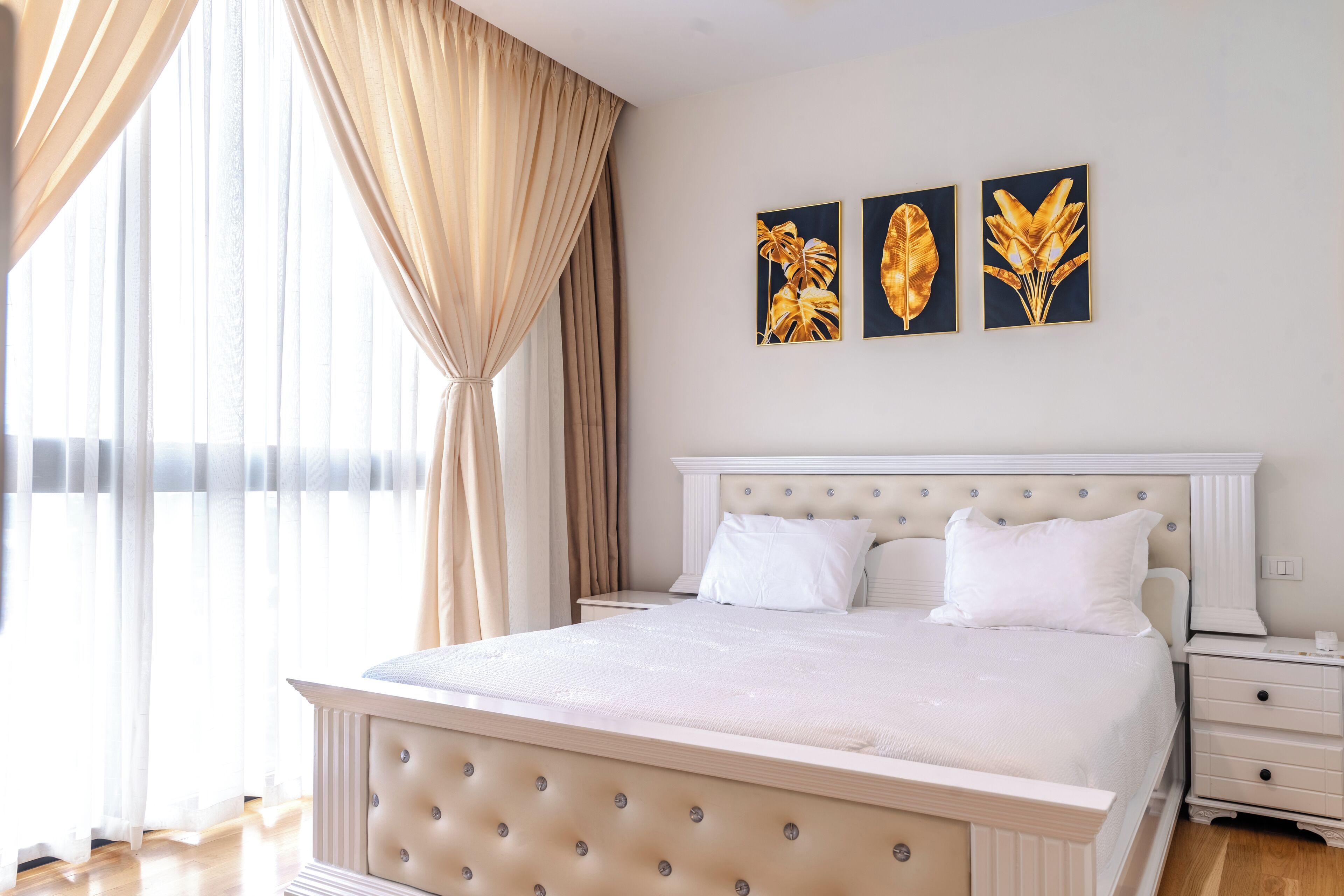 A bedroom with a large bed, a nightstand, and a window with curtains. There are three framed pictures on the wall.