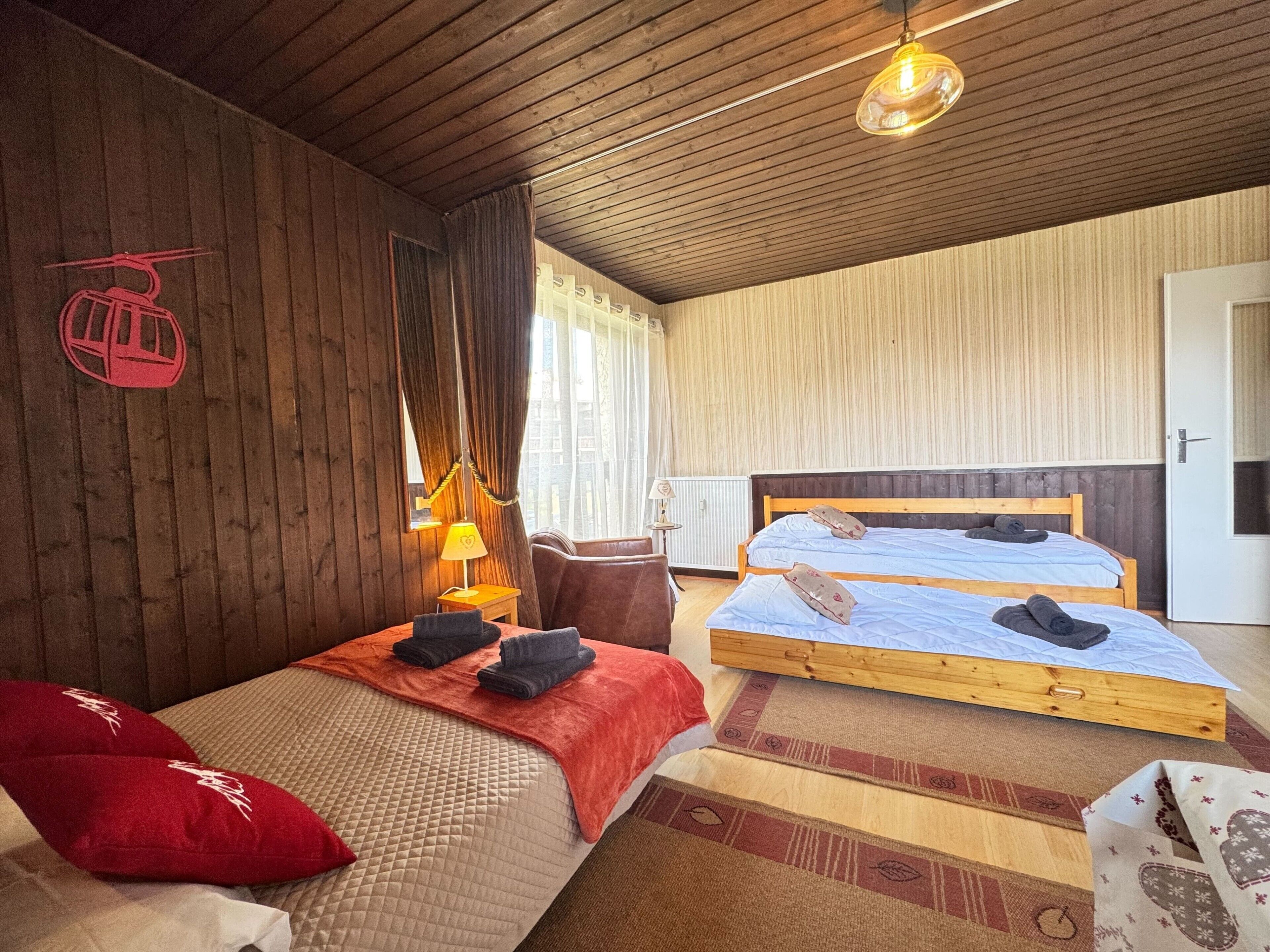 A room with two wooden beds, a red chair, a small table with a lamp, and a wooden ceiling.