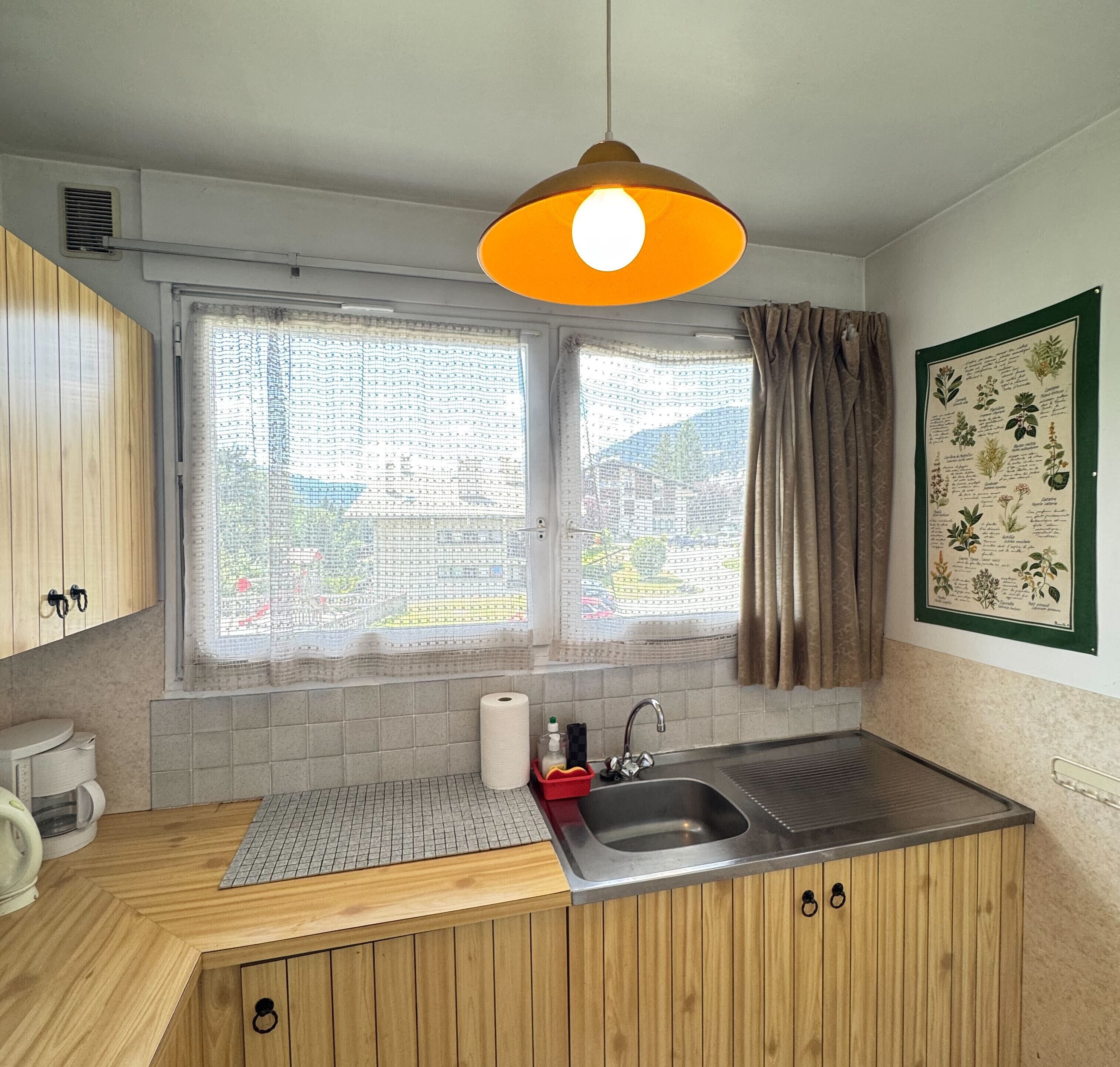 Een keuken met houten kastjes, een roestvrijstalen spoelbak, een raam met gordijn en een hanglamp.
