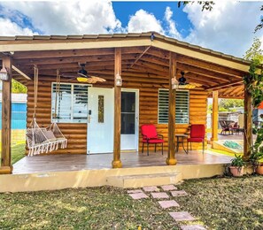 Una cabaña de troncos con un porche cubierto, una hamaca, sillas rojas y una zona de piscina.