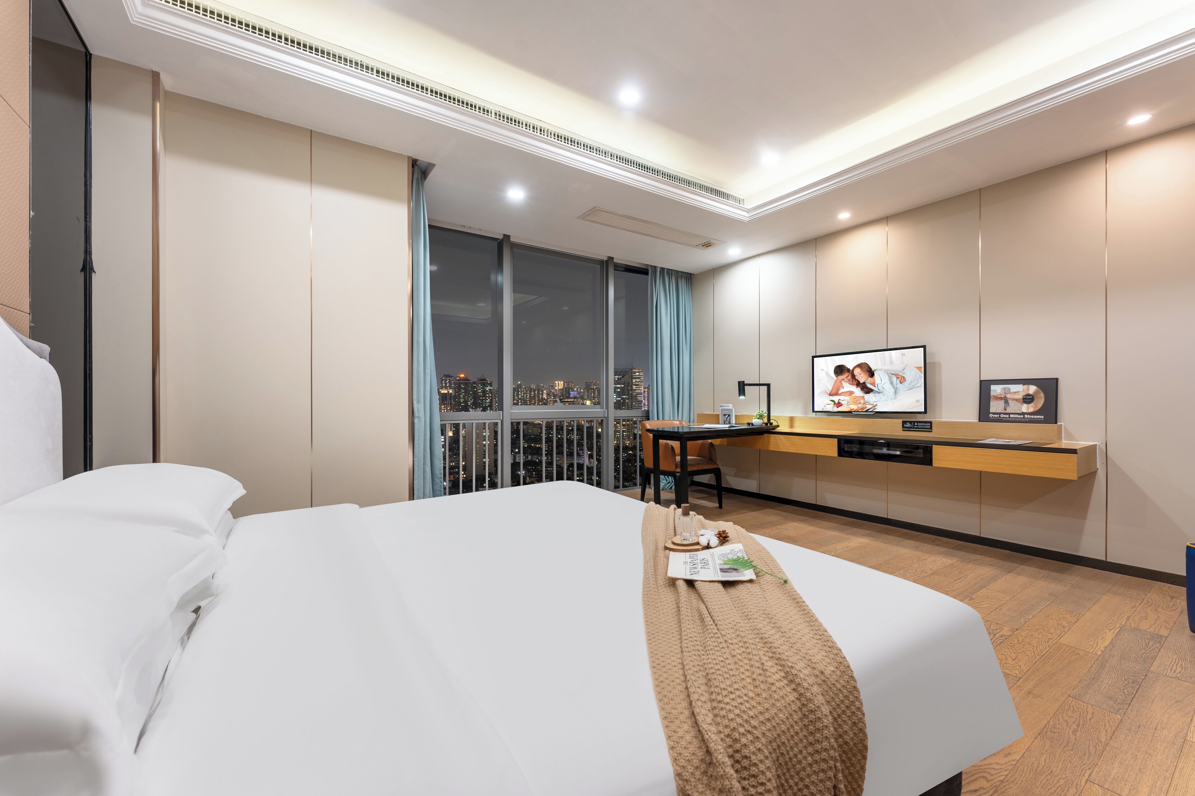 A hotel room with a bed, a desk with a chair, a television, and a city view through large windows.