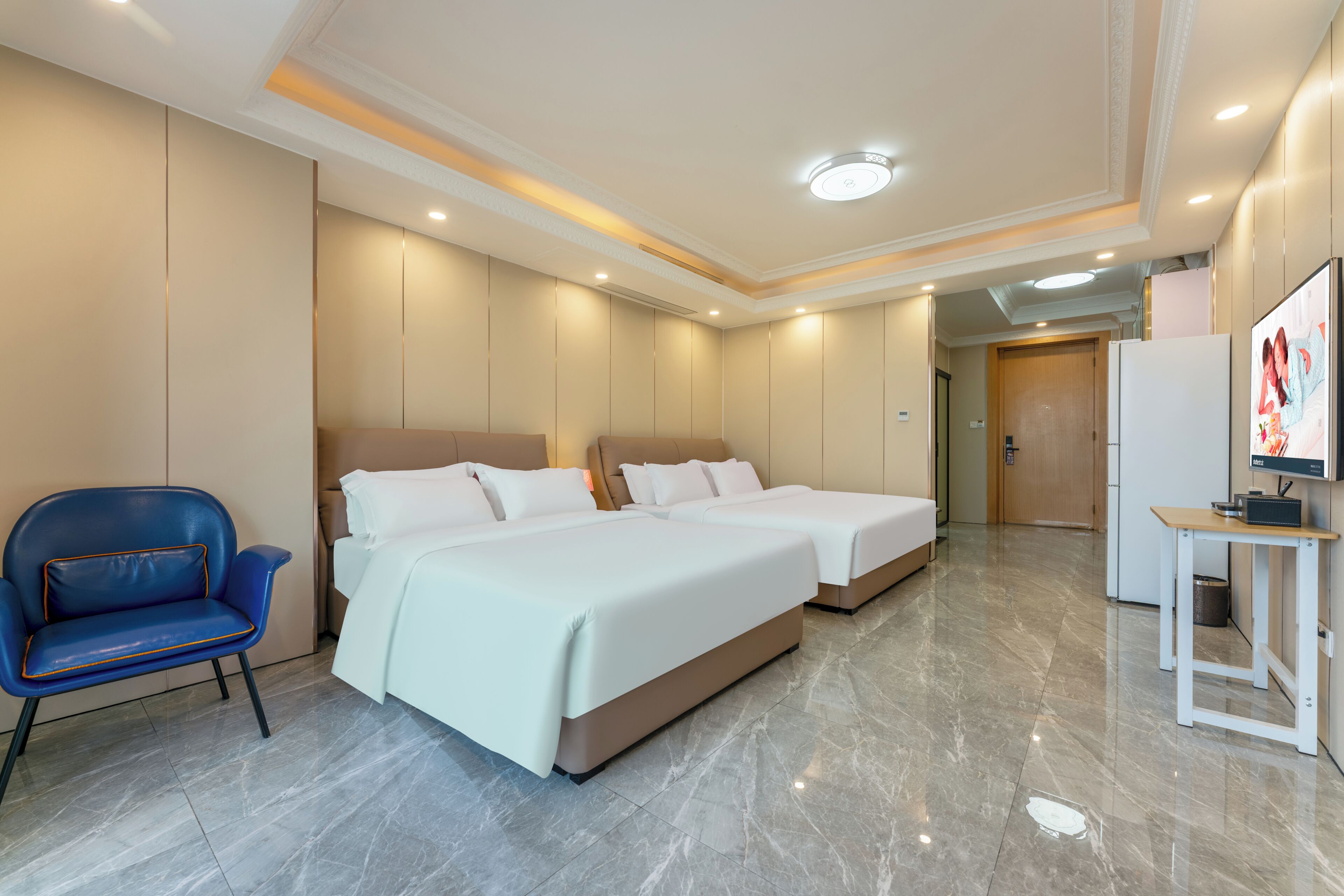 A hotel room with two beds, a blue chair, a small table, and a door leading to another room.