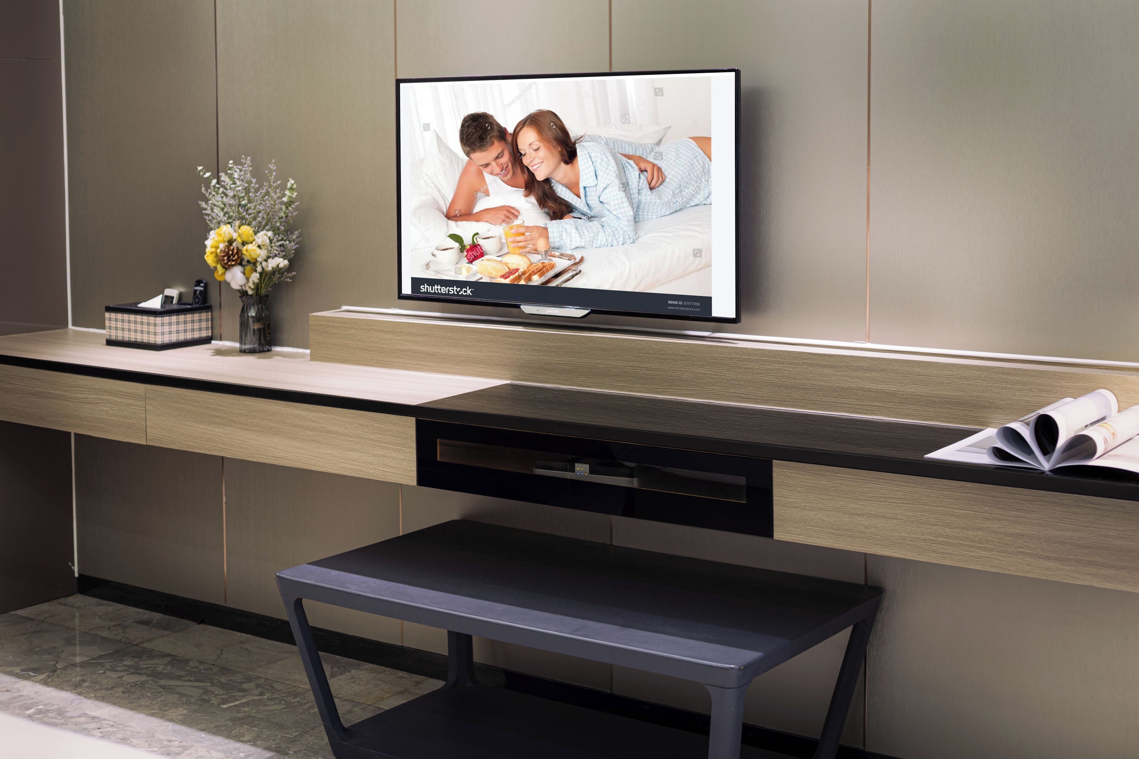 A modern hotel room with a flat-screen TV mounted on a wall, a wooden desk with a bench, and a vase with flowers on the side.