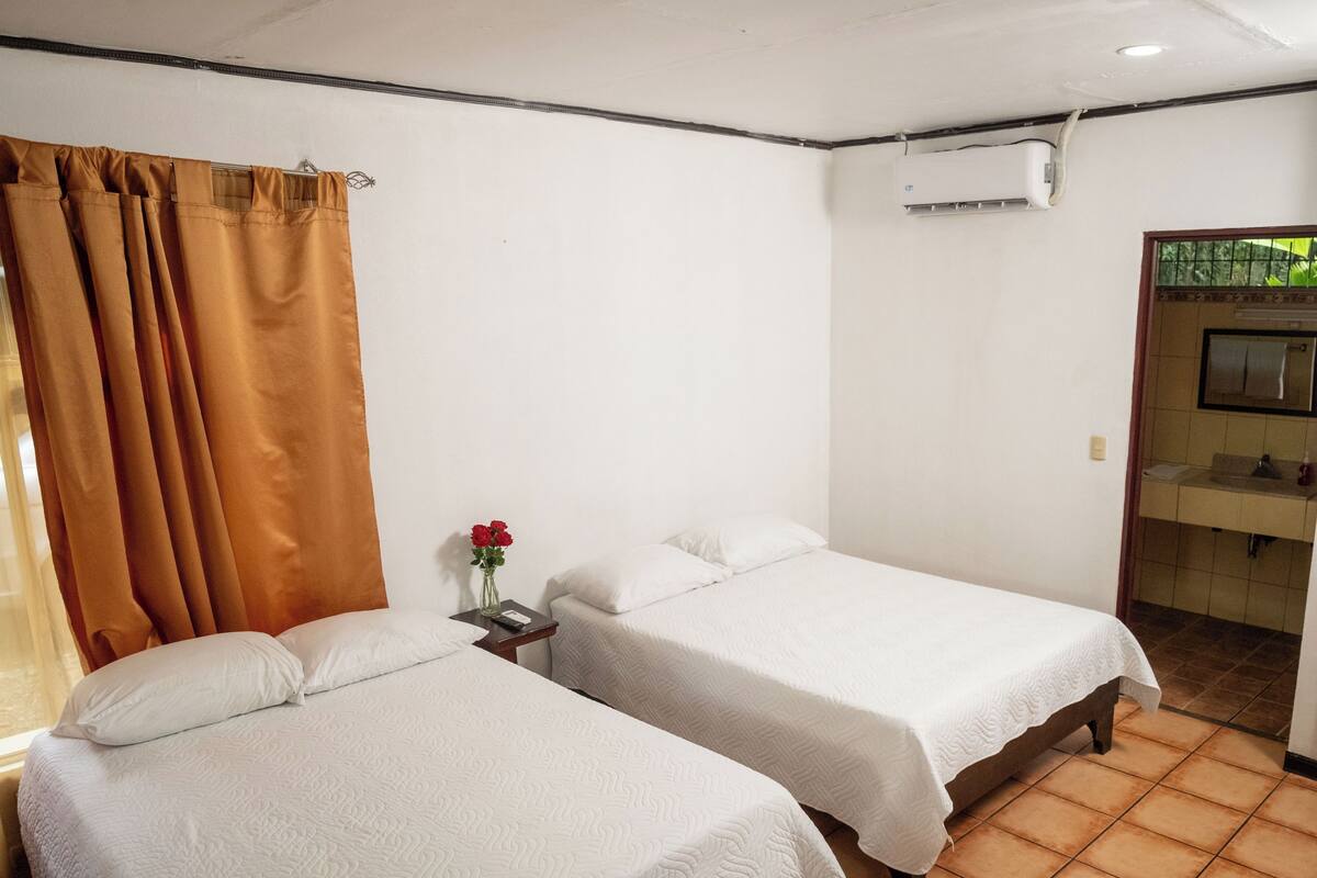 Een hotelkamer met twee bedden, een nachtkastje met een vaas rode bloemen, een badkamer met een wastafel en spiegel, en een airconditioningunit aan de muur.
