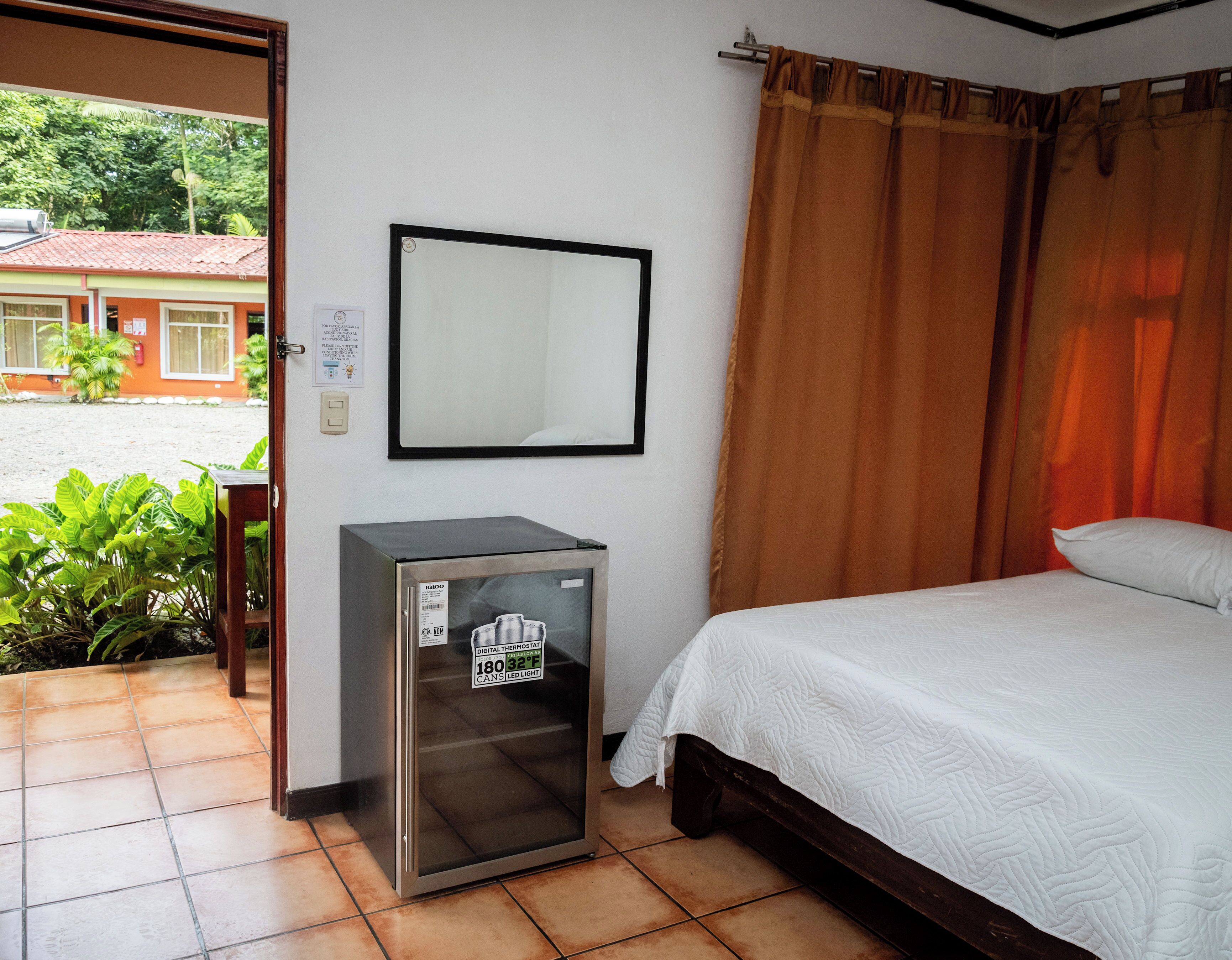 Quarto de hotel com cama, pequeno frigorífico, espelho e vista para o exterior.