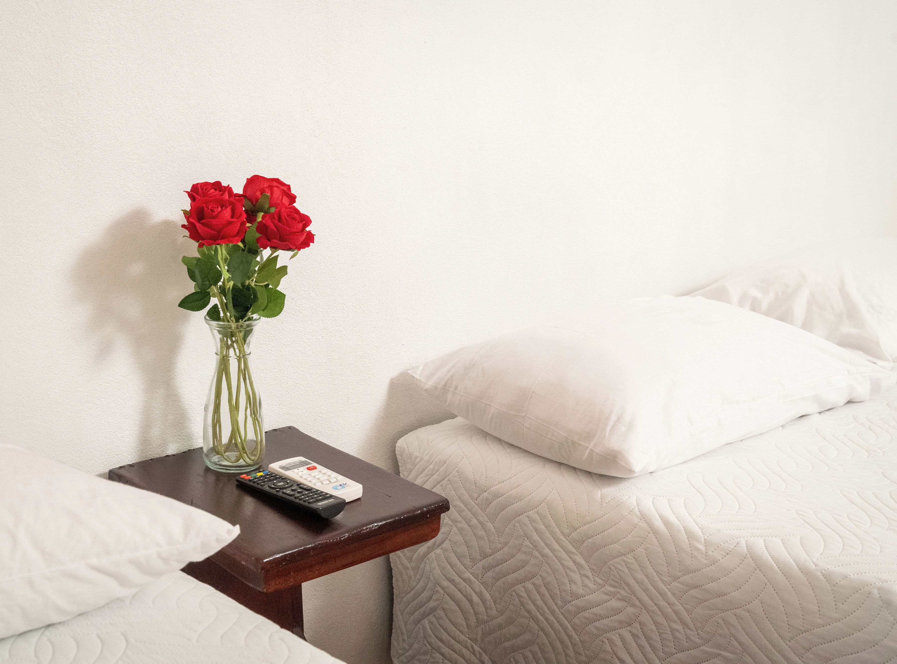 Quarto de hotel com cama, almofadas brancas, mesa de cabeceira com um vaso de rosas vermelhas e controle remoto.