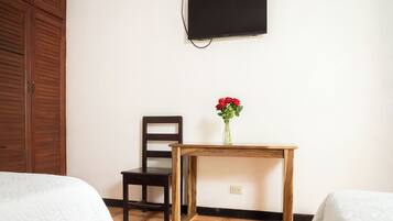 A room with a bed, a wooden chair, a small wooden table with a vase of red flowers, and a flat-screen TV mounted on the wall.