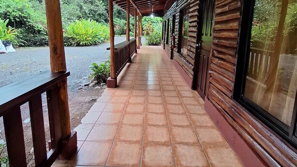 A covered walkway with wooden pillars and a tiled floor, leading to a building with large windows. - Hotel Sun Sun Lodge (Horquetas)