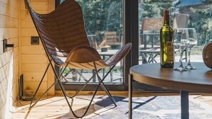 A modern interior with a wooden ceiling, a large window, a folding chair, a table with a bottle and glass, and a view of trees and buildings outside. - Mountain Home Durmitor 1 (Uskoci)