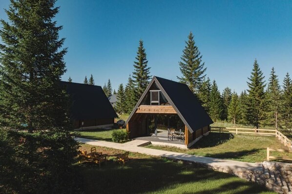 Une cabane dotée d’un toit triangulaire et d’un porche, entourée d’un mur de pierres et d’une clôture en bois, située dans une zone boisée.