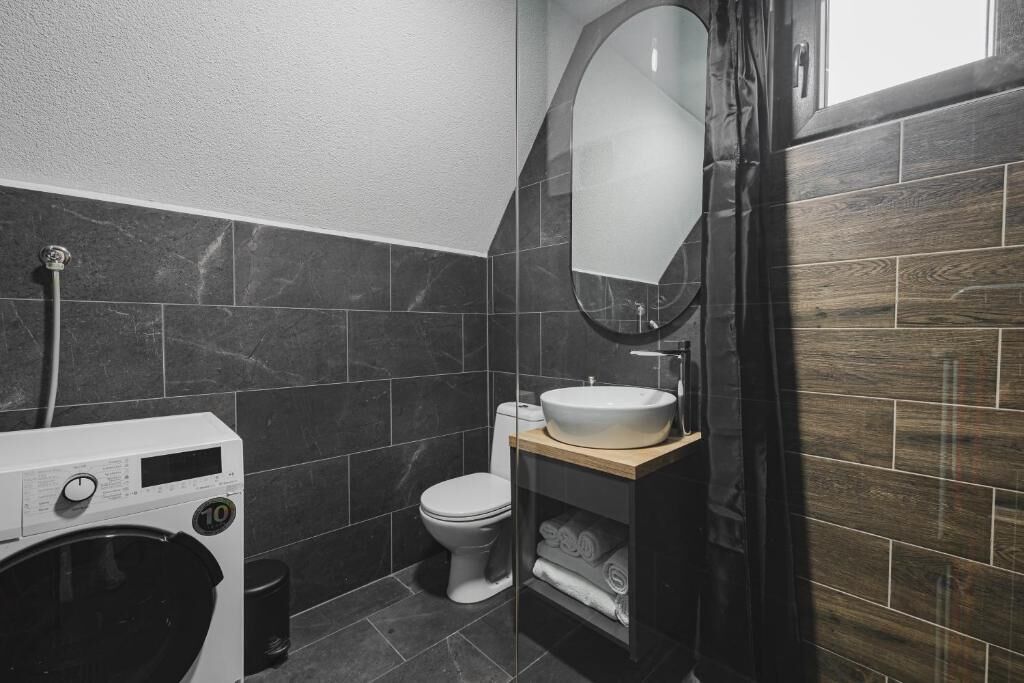 Een kleine badkamer met een wasmachine, toilet en een houten wastafelmeubel met spiegel.