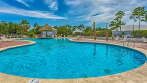A clear blue swimming pool with lounge chairs and a thatched-roof hut, surrounded by trees and a clear sky.