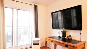 A room with a large window, a standing heater, a wooden desk with a flat-screen TV, and a small plant on the desk.