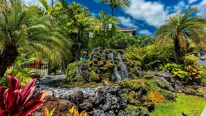 Un jardín tropical con una cascada, abundante vegetación y una casa al fondo.