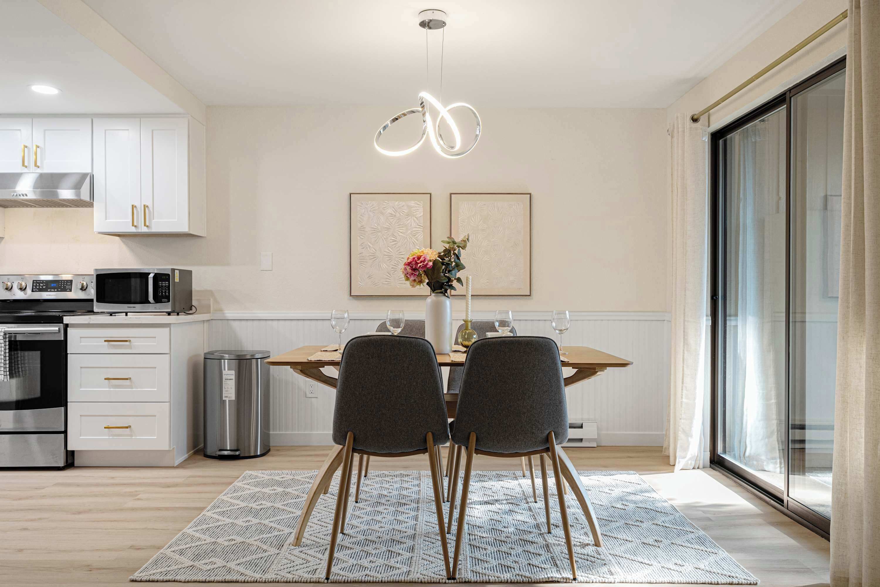 Una cocina moderna con armarios blancos, electrodomésticos de acero inoxidable y un comedor con mesa de madera y dos sillas.