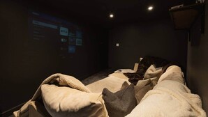 A dimly lit room with a bed covered in blankets, a wall-mounted projector displaying a menu, and a small table with a lamp.