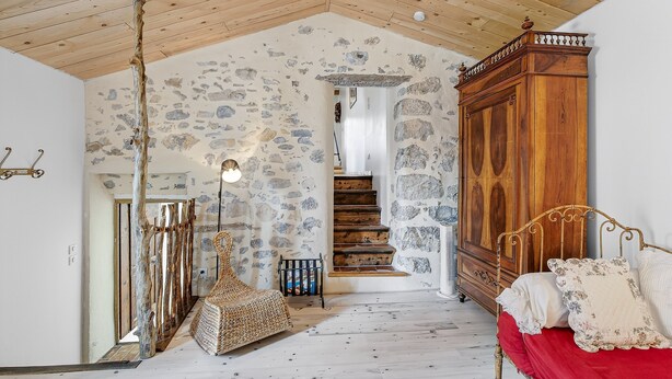 Ein rustikaler Raum mit Steinwand, Holzboden, einem Bett mit roter Bettwäsche, einem hölzernen Kleiderschrank und einer Treppe, die zu einer weiteren Ebene führt.