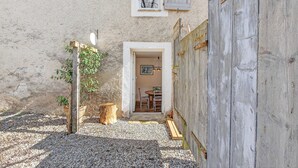 Ein rustikaler Außenbereich mit Kiesweg, Holzlattenzaun und einer Tür, die zu einem Innenraum mit Tisch und Stühlen führt.