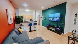 A modern living room with a grey sofa, two blue bar stools, a TV on a teal accent wall, and a kitchen in the background. - Convention Cent / 2 King Beds / 65" TV / Fireplace (Philadelphia)