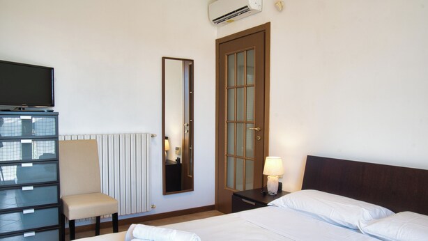 A hotel room with a bed, a chair, a desk with a flat-screen TV, a mirror, a door, and a lamp.