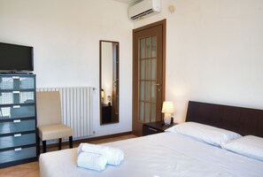 Habitación de hotel con cama, una silla, escritorio con televisor de pantalla plana, espejo, puerta y lámpara.