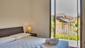 A hotel room with a bed, a nightstand, a lamp, and a balcony with a view of buildings and greenery.