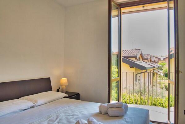 Habitación de hotel con cama, mesita de noche, lámpara y balcón con vistas a edificios y vegetación.