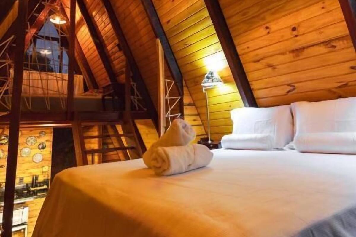 A cozy attic bedroom with a wooden ceiling, a bed with white linens, and a hanging light fixture.