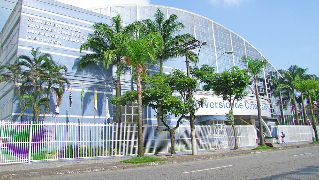 A modern building with a curved glass facade, surrounded by a white fence and palm trees.