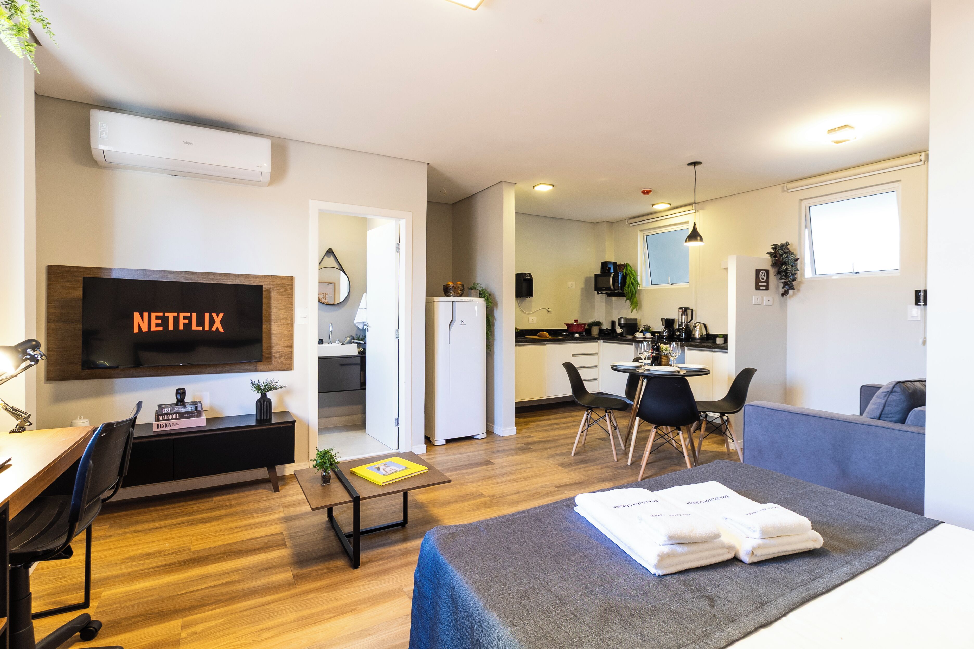 A modern hotel room with a flat-screen TV, a desk, a sofa, a dining area, and a kitchenette.