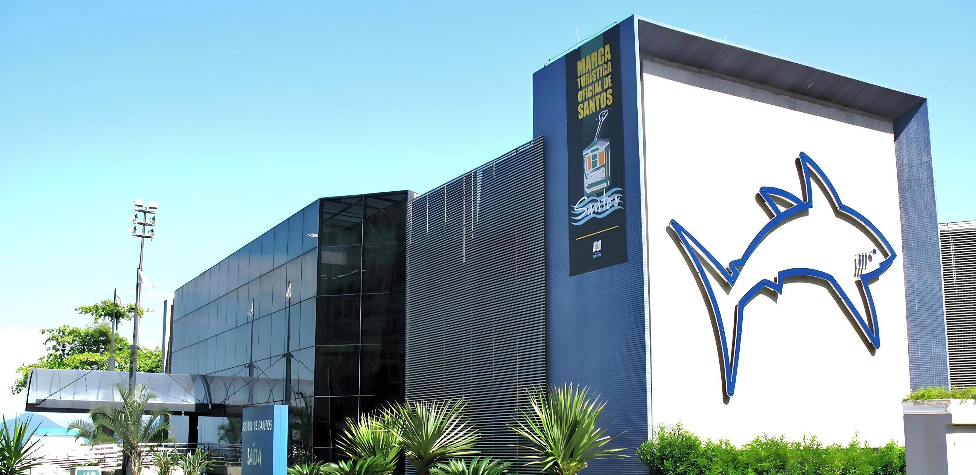 A modern building with a large blue logo, a sign for 'MARCA TOURISTA ORICIAL DE SANTOS,' and a covered area with plants.
