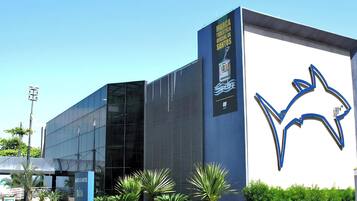 A modern building with a large blue logo, a sign for 'MARCA TOURISTA ORICIAL DE SANTOS,' and a covered area with plants.