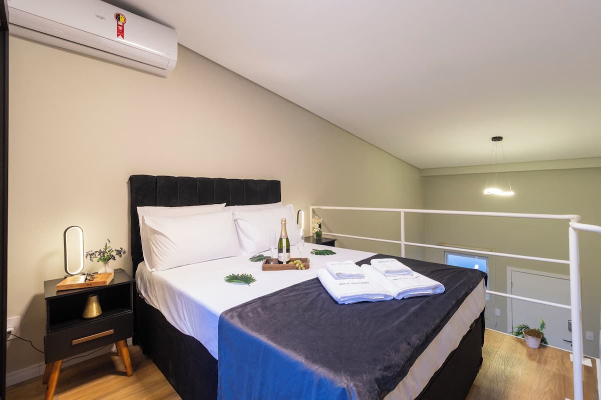 Een netjes opgemaakt bed met een zwart hoofdbord, een nachtkastje met een spiegel, een kleine plant en een dienblad met een fles en druiven.