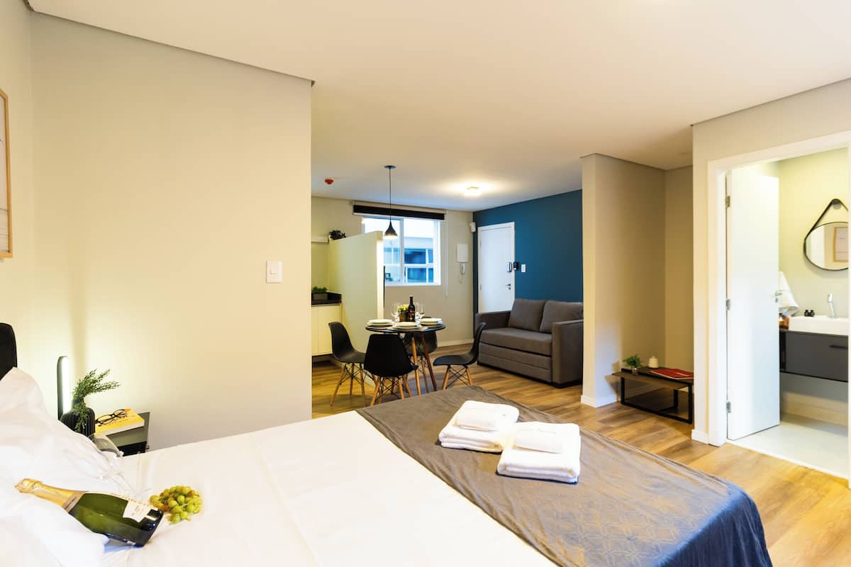 Hotelkamer met een bed, een eettafel met stoelen, een bank, een bijzettafeltje met een lamp, en een zichtbare badkamer door een open deur.