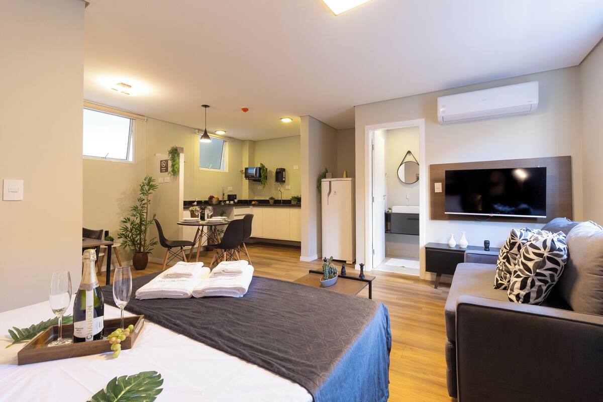 Een moderne hotelkamer met een bed, een televisie, een zithoek en een keuken op de achtergrond.