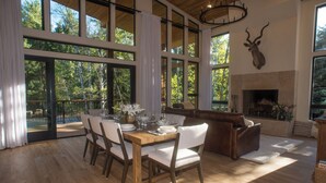 A spacious dining area with a wooden table and chairs, large windows offering views of trees, and a stone fireplace. - Beautiful Mountain Modern Home with well equipped kitchen. Perfect for families. (Ketchum)