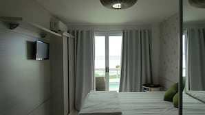Habitación de hotel con cama, televisor, balcón con cortinas y vista al mar.