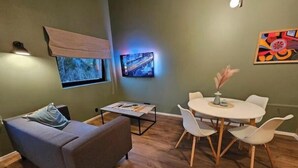A modern living room with a grey sofa, a small table, a dining table with chairs, a TV mounted on the wall, and a window with a view of a bridge.