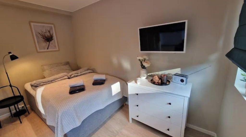 A bedroom with a bed, bedside table, lamp, and a flat-screen TV mounted on the wall.