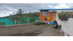 Una pequeña construcción de madera con techo azul, una motocicleta estacionada adelante y un área de jardín con plantas y una cerca.