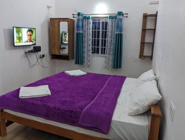 Un dormitorio con cama de madera, ropa de cama color morado, televisor montado en la pared, ventana con cortinas y una pequeña estantería de madera.