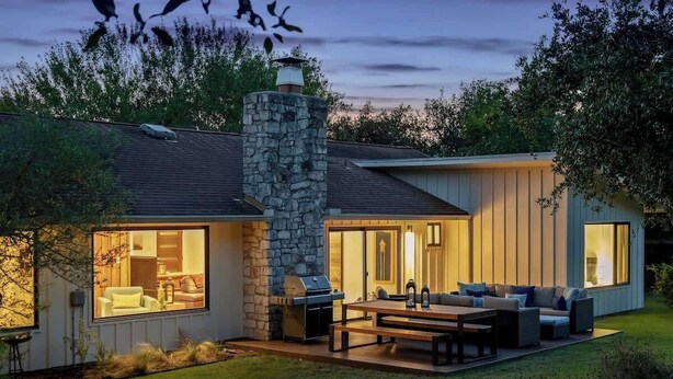 A modern house with a stone chimney, large windows, and an outdoor seating area with a table and chairs. - Nestled In Nature Home - Pet-Friendly, Acreage (Austin)