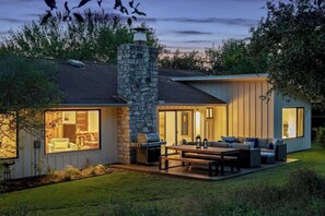 A modern house with a stone chimney, large windows, and an outdoor seating area with a table and chairs. - Nestled In Nature Home - Pet-Friendly, Acreage (Austin)